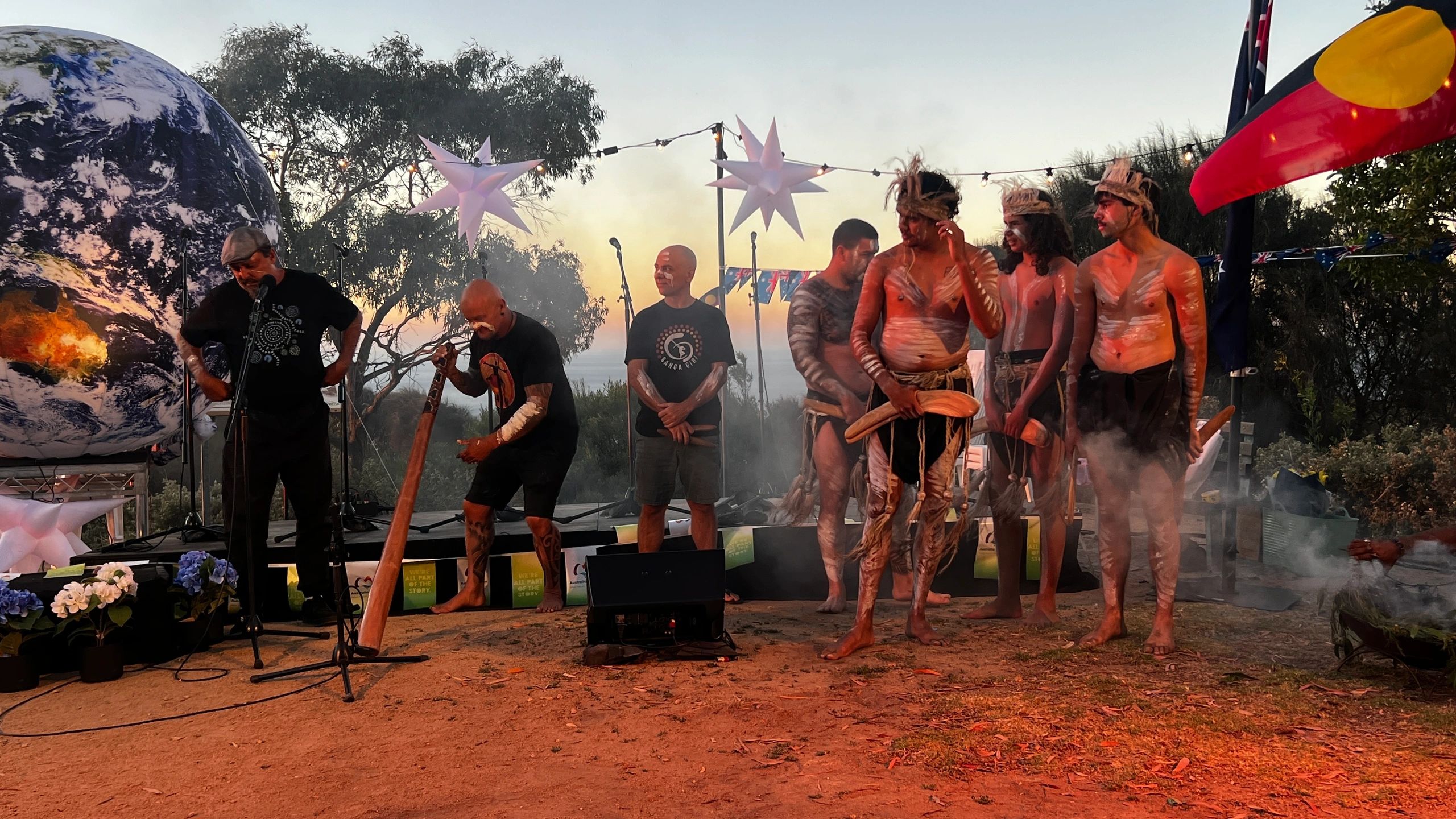 Cultural ceremony at dusk with performers, smoke, and coastal light